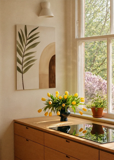 Akustikbillede med et grønt palmeblad ved en terrakottafarvet bue, ved et vindue med en blomsterbuket i karmen