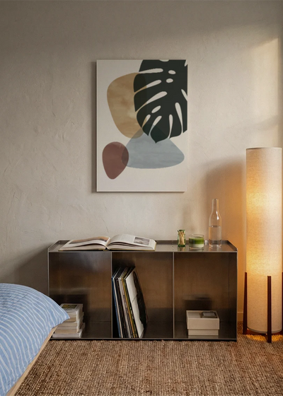 Akustikbillede med et sort monsterablad med beige og grå former, over sengegavlen i et soveværelse med tændt lampe