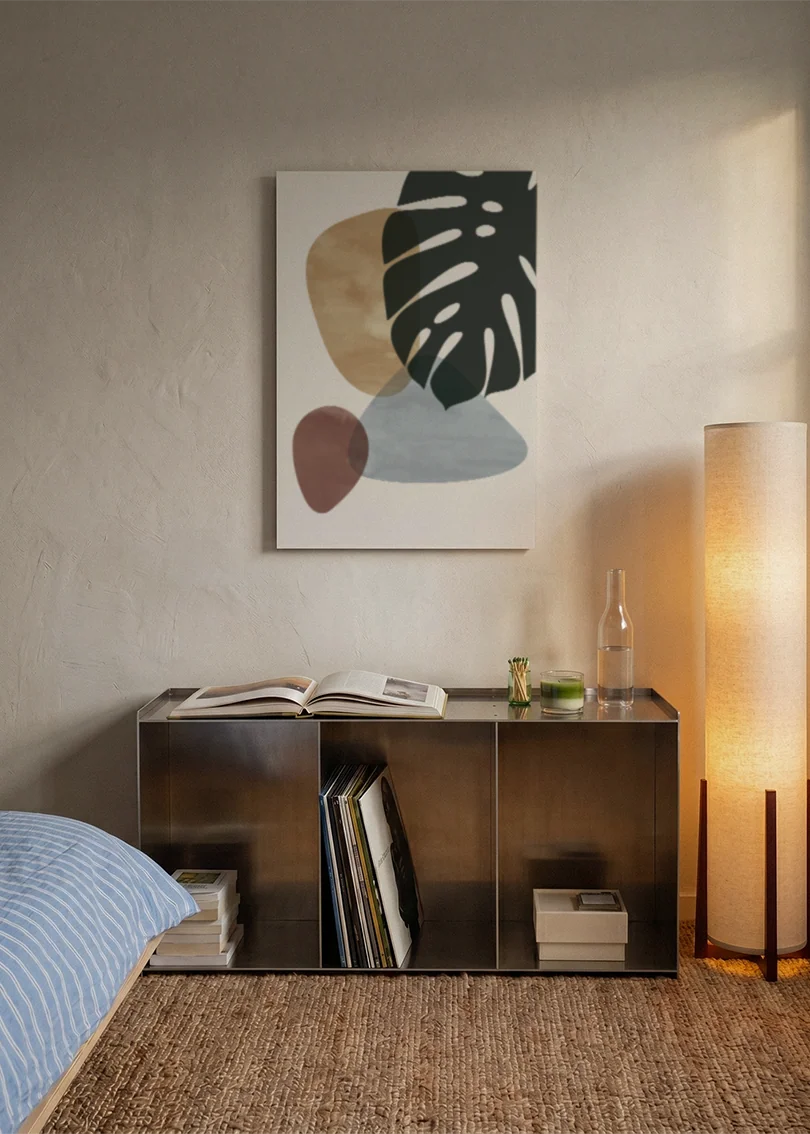 Akustikbillede med et sort monsterablad med beige og grå former, over sengegavlen i et soveværelse med tændt lampe