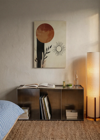 Akustikbillede med en terrakottacirkel med en lille sol og sorte former, over sengegavlen i et soveværelse med tændt lampe