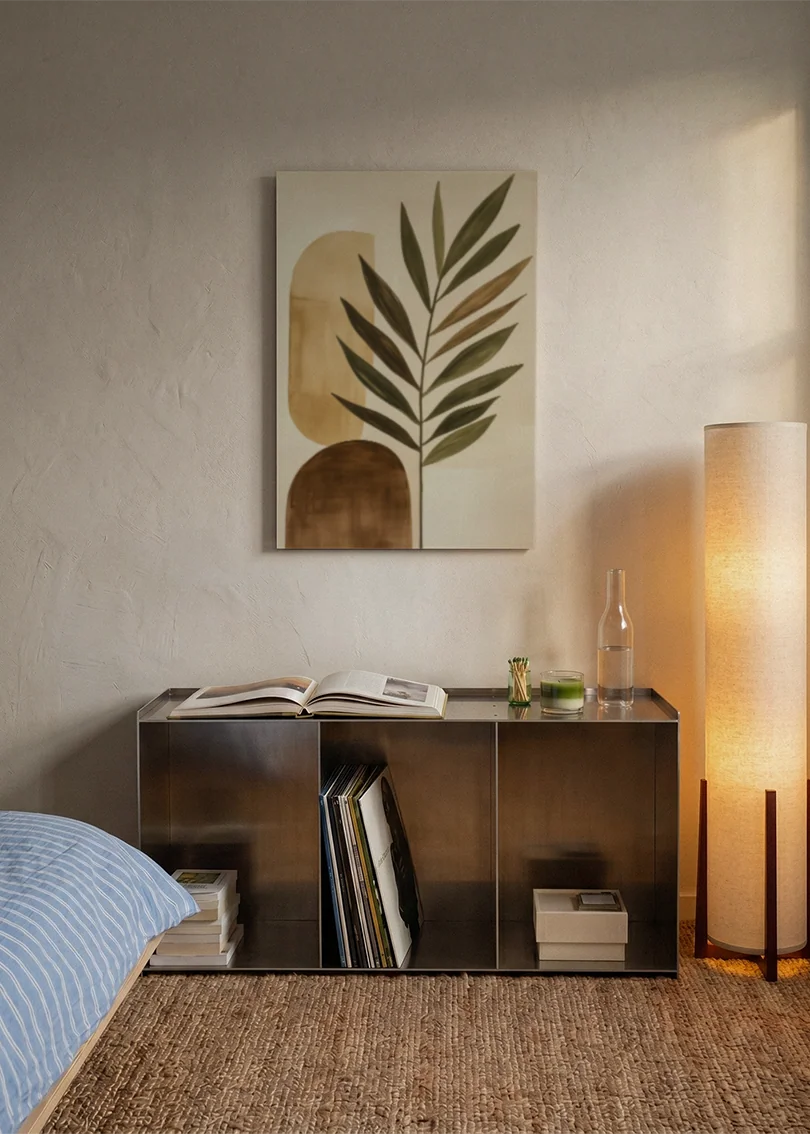 Akustikbillede med et stort grønt blad med beige former, over sengegavlen i et soveværelse med tændt lampe