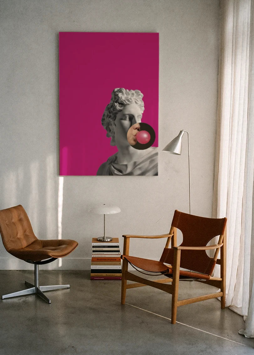 Akustikbillede med en gipsbuste med en tyggegummiboble på pink baggrund, ophængt over en lænestol ved gardinet med gulvlampe