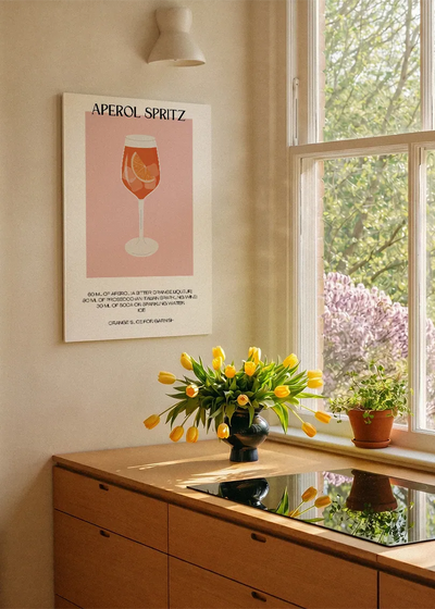 Akustikbillede med en minimalistisk cocktailplakat med Aperol Spritz, ved et køkkenvindue med gule tulipaner