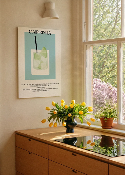 Akustikbillede med en minimalistisk cocktailplakat med Caipirinha, ved et køkkenvindue med gule tulipaner