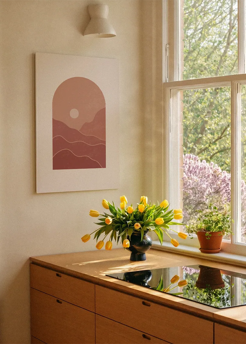 Akustikbillede med rosa bakker med en sol i en bueform, ved et vindue over køkkenbordet med gule tulipaner