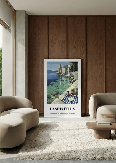 Akustikbillede med en italiensk rejseplakat med hvide klipper ved Middelhavet, i en lounge med bugtet sofa mod vægpanel i valnød