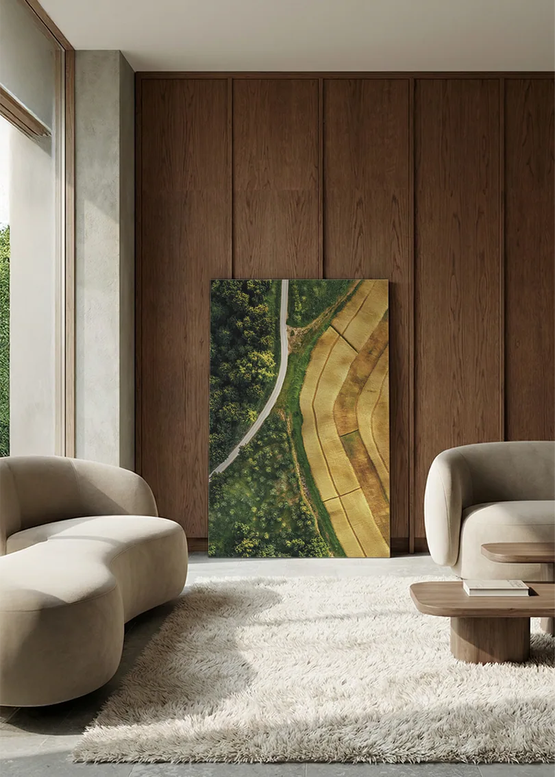 Akustikbillede med dronefoto af en markvej mellem grøn skov og gyldne marker, i lounge med bugtet sofa mod vægpanel i valnød