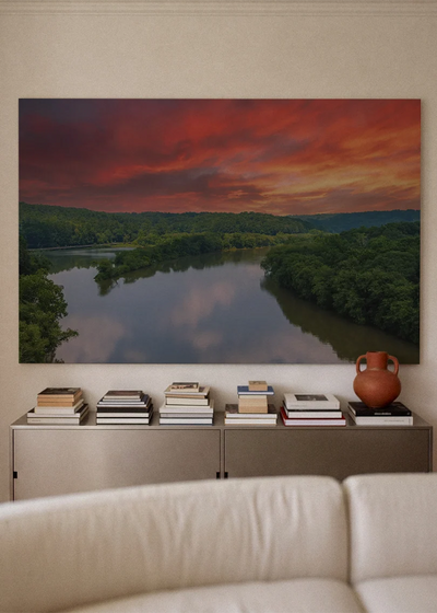 Akustikbillede med en flod ved solnedgang under rød og orange himmel, over sofa med bøger og terrakottavase