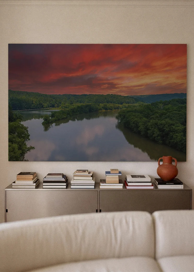 Akustikbillede med en flod ved solnedgang under rød og orange himmel, over sofa med bøger og terrakottavase
